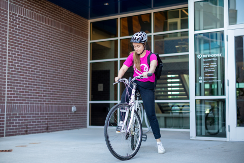 Woman in pink Methodist Health System shirt getting on a bike in front of a brick building.